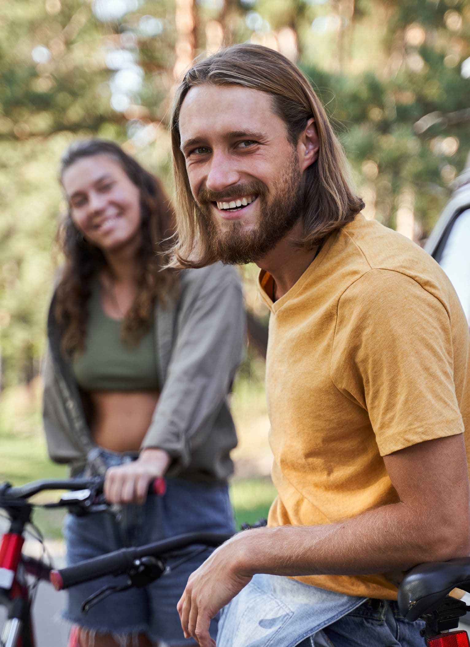 A young man and woman enjoying a day outdoors. The man, with shoulder-length hair and a beard, smiles towards the camera while sitting on a bicycle. The woman stands slightly behind on another bike, smiling in the background. The scene is relaxed and sunny, with soft-focus trees behind them.