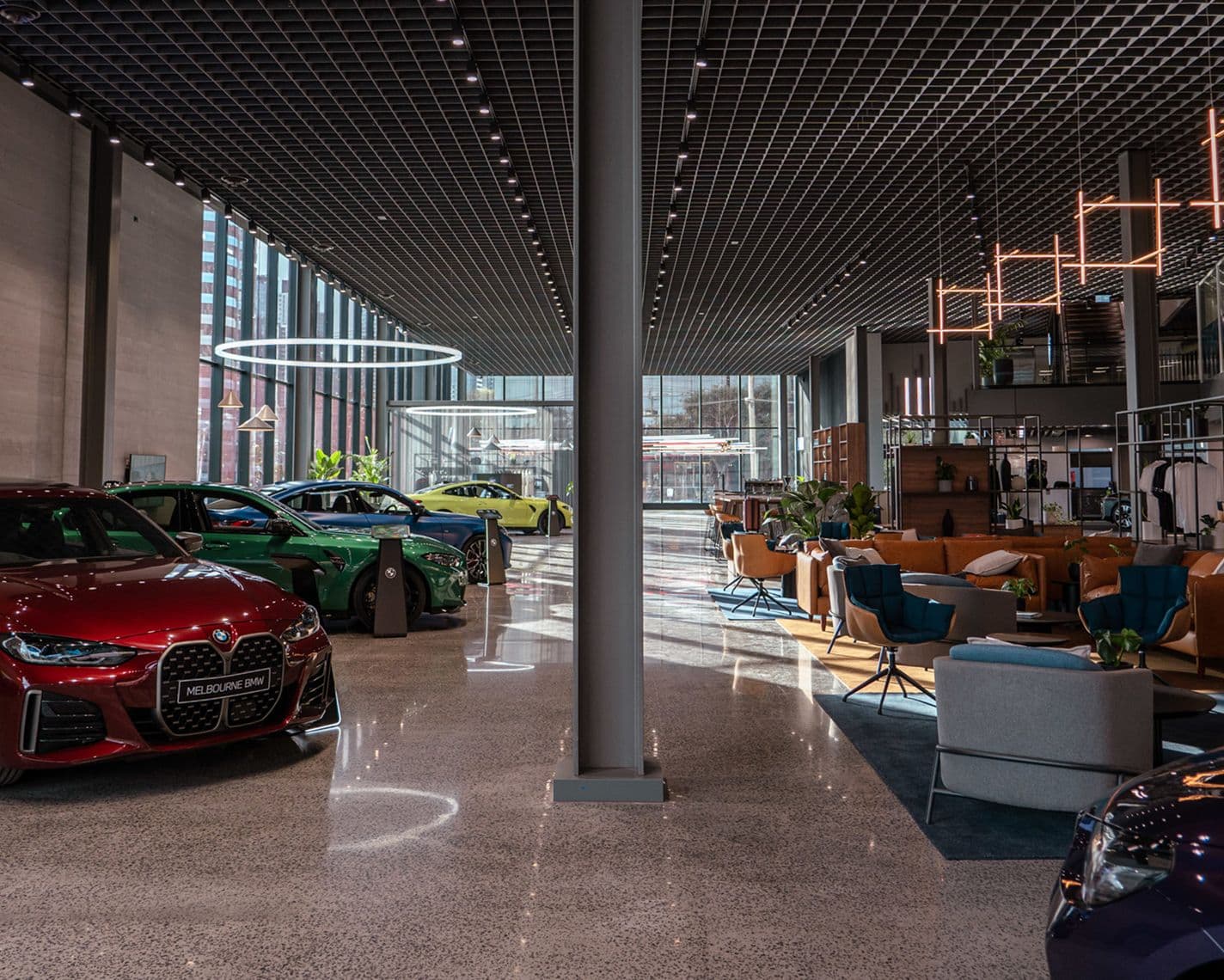 "Modern BMW showroom with several luxury cars on display, including a red, green, and yellow model. The space features high ceilings with grid lighting, large glass windows letting in natural light, and a stylish lounge area with contemporary seating and plants.