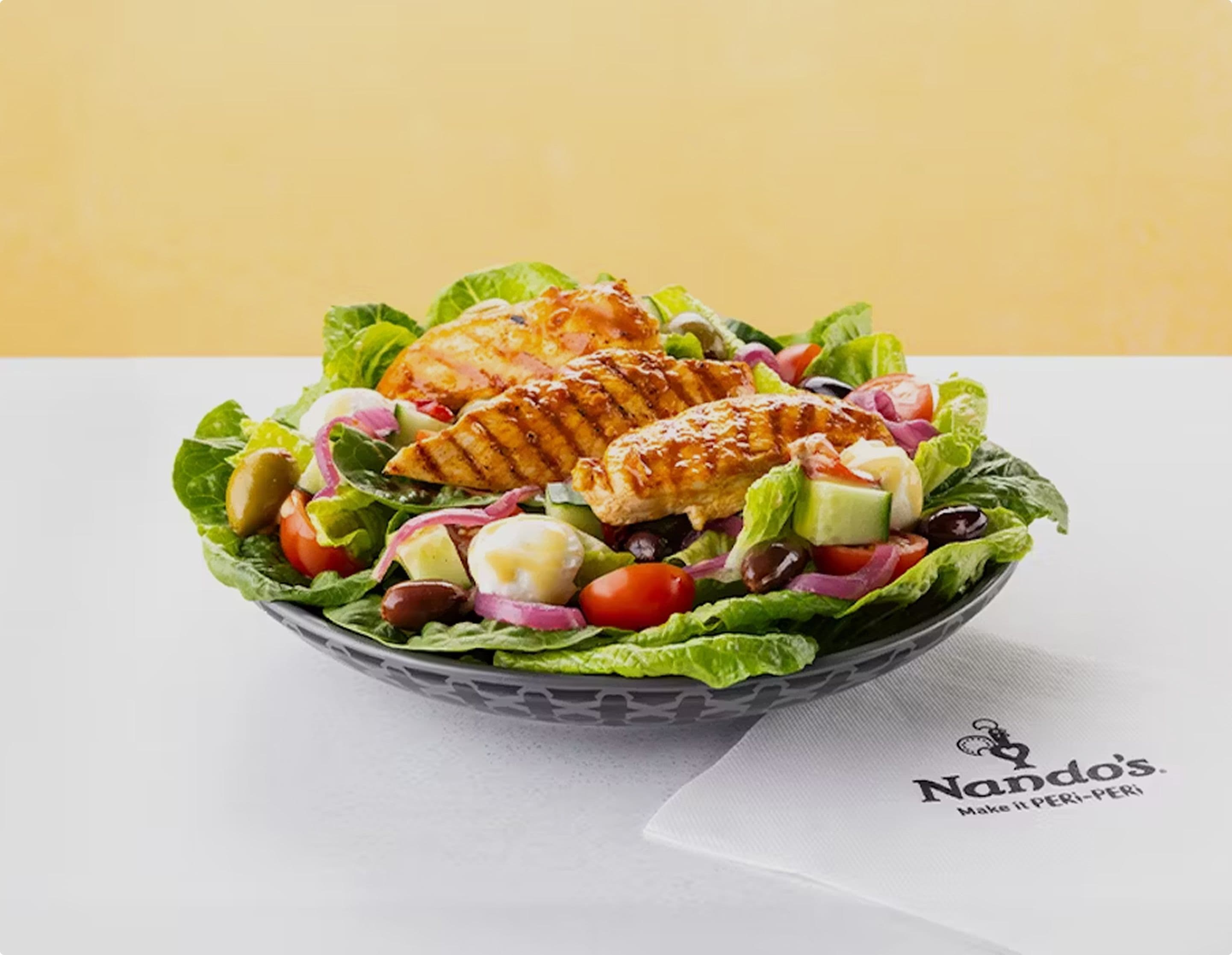 Plate of fresh salad topped with grilled chicken, cherry tomatoes, cucumber, olives, feta cheese and red onion, placed on a white table with a Nando’s branded napkin.