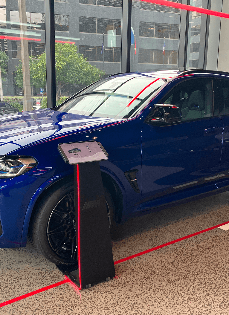 Blue BMW X6 M Competition SUV displayed inside a modern showroom, positioned next to a sleek black and red information stand. The car’s front half is visible, with sunlight reflecting off its glossy surface and city buildings in the background.