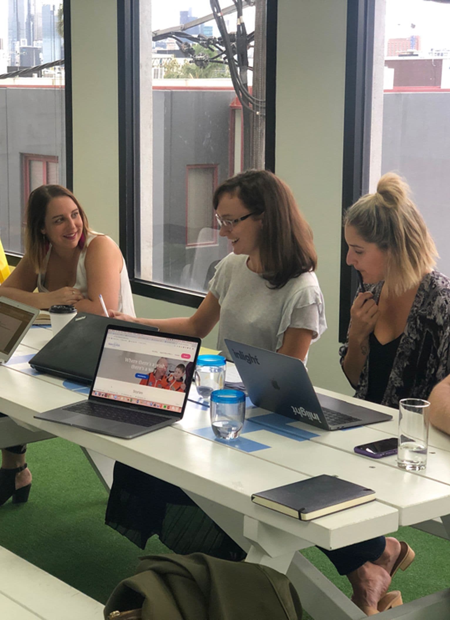 People are gathered around a white table in a modern office, collaborating on laptops and taking notes. Branded water bottles and notebooks are visible on the table. Large windows in the background show city buildings, and the workspace feels bright and open. One laptop displays a homepage for Make-A-Wish Australia.