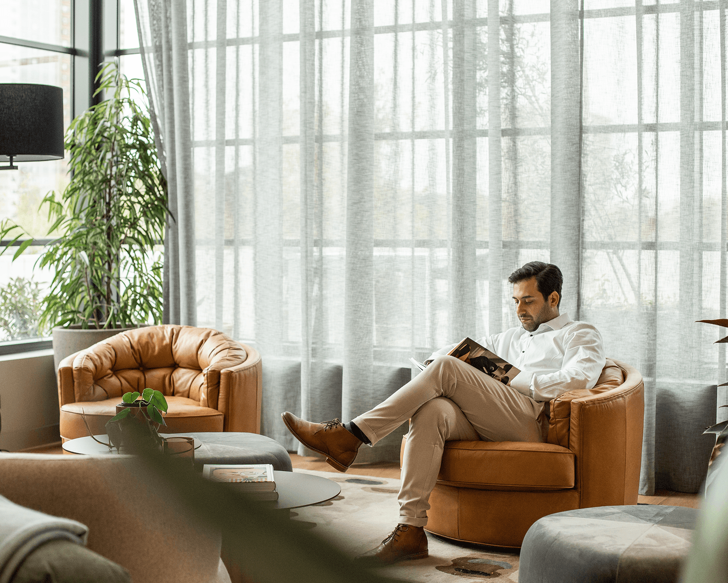 A serene lounge space with large windows covered by sheer curtains, filling the room with natural light. Comfortable leather armchairs and round tables are arranged for relaxation, alongside potted plants. A person sits reading a magazine, dressed business-casual, creating a calm and studious atmosphere.