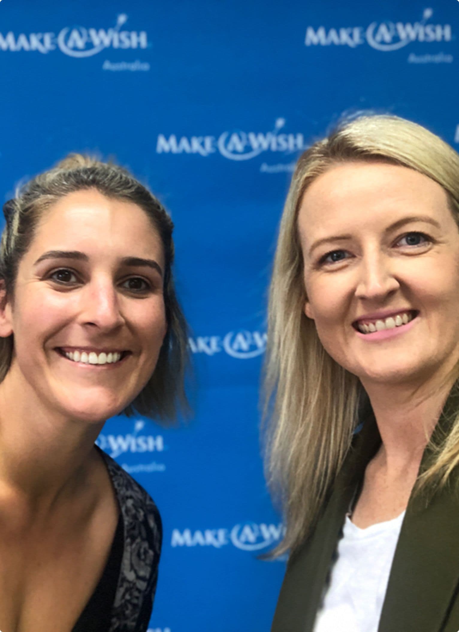 Two people stand in front of a blue Make-A-Wish Australia media wall, which features the charity’s white logo repeated across the backdrop. The setting suggests a friendly event or partnership showcase, with an atmosphere of positivity and connection.