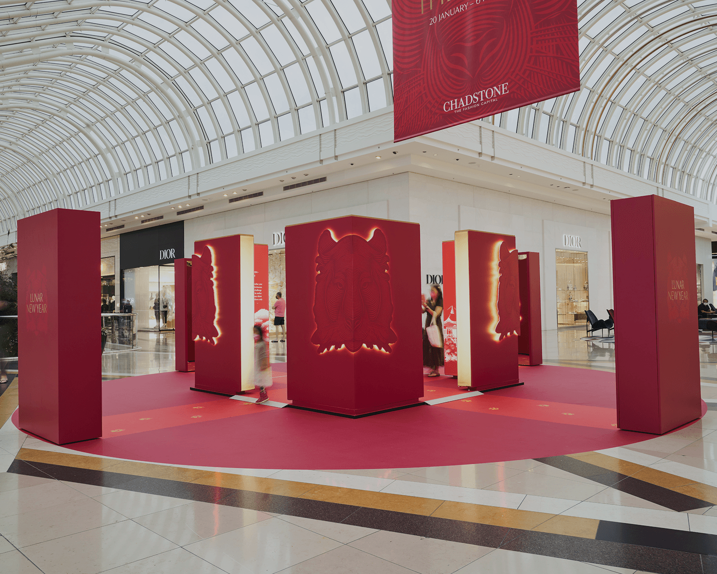 A modern shopping centre atrium with a curved glass roof. Large red Lunar New Year installations displaying tiger motifs are arranged in a circle beneath a prominent event banner and surrounded by luxury stores such as Dior.