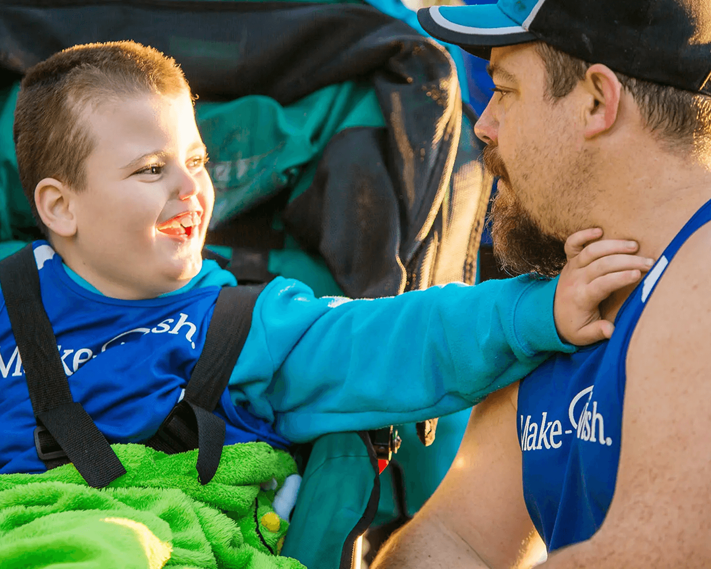 A smiling young boy in a wheelchair reaches out to a bearded man in a Make-A-Wish shirt. They are sharing a joyful moment outdoors, showing connection and support.