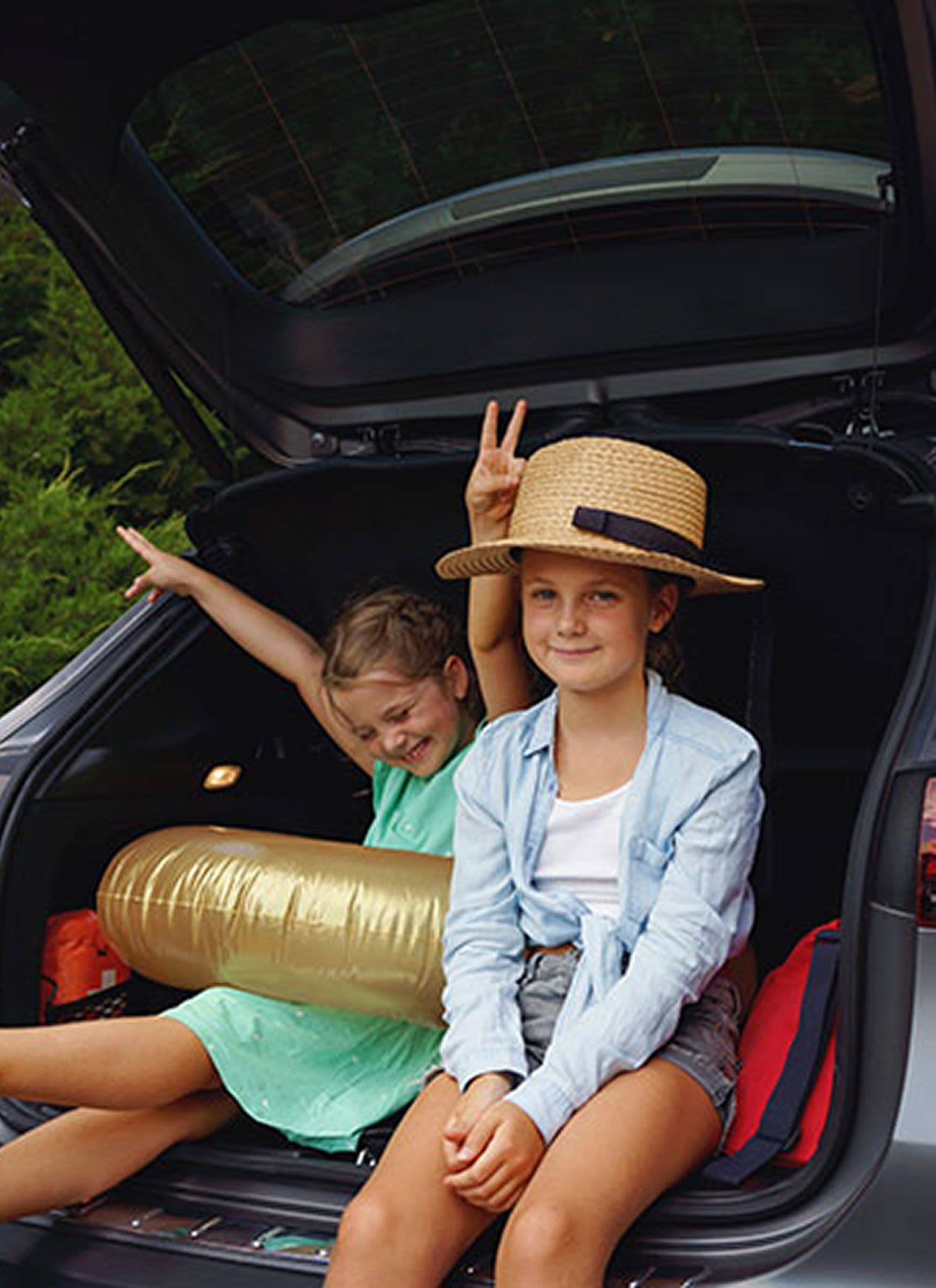 Two children sitting in the open boot of a car, one wearing a straw hat and light blue shirt, and the other in a green dress holding a gold inflatable pool toy, with green foliage in the background.