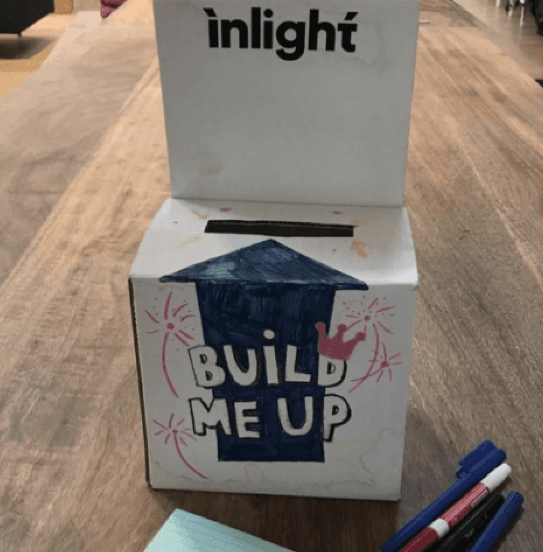 Cardboard box labelled “Build Me Up,” decorated with colourful designs and pens and paper nearby, designed for collecting positive feedback and encouragement from team members.