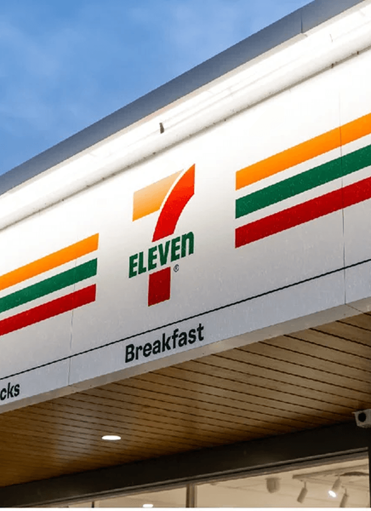 The exterior sign of a 7-Eleven store with its orange, green, and red stripes and logo above the entrance.