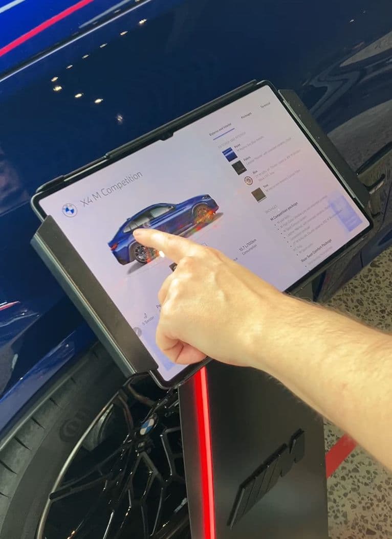 a hand pointing at a tablet screen that is next to a car inside a BMW dealership. The screen has an image of the car and specifications about the car
