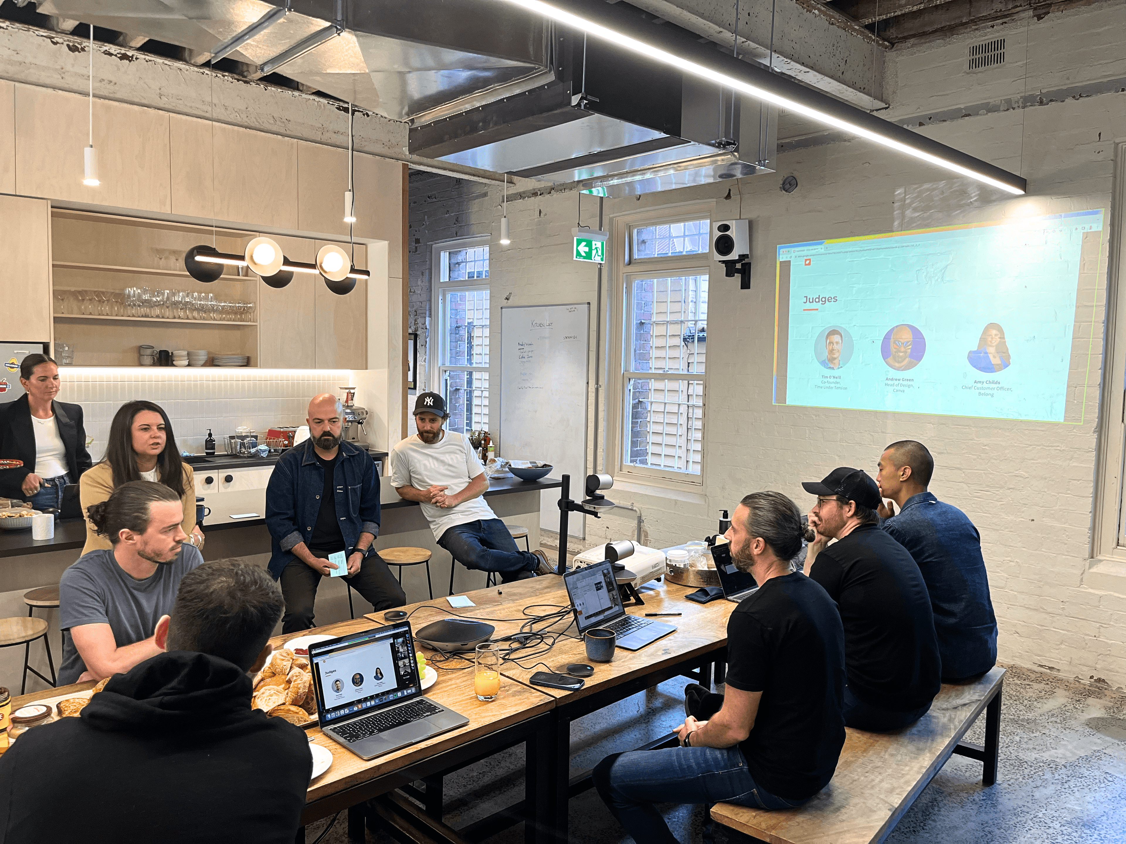 A group of colleagues gathers in an open kitchen workspace, watching a presentation projected onto a wall. Some sit at a long table with laptops and refreshments, while others stand or sit at the bench, engaged in the session.