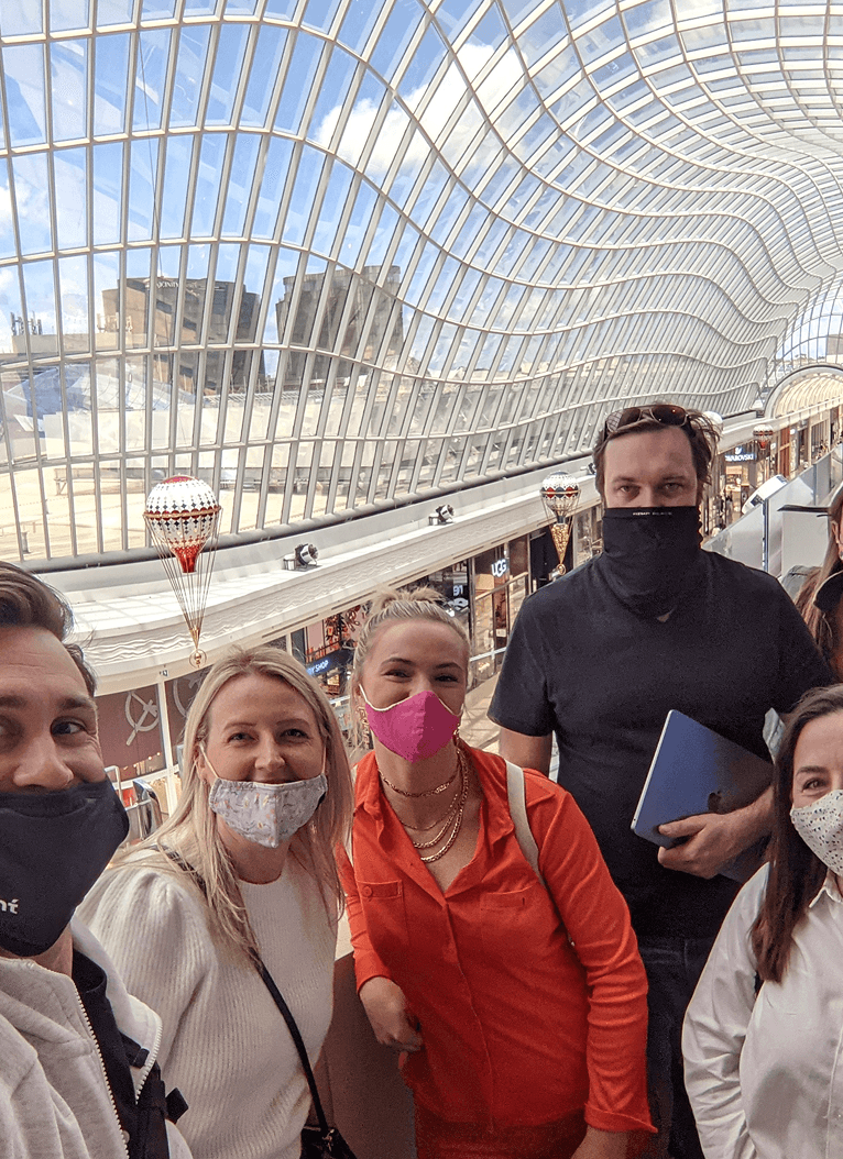 A group of people stand inside a shopping centre under a large, curved glass roof. Behind them, a row of shops is visible under natural daylight, with festive decorations hanging from the ceiling.
